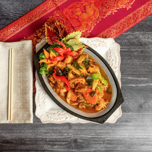 Sizzling platter of crispy chicken with vegetables and sauce, chopsticks on a napkin, on a dark wooden table.