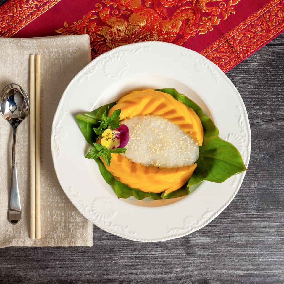Mango sticky rice dessert on a white plate with a spoon and chopsticks on a napkin.