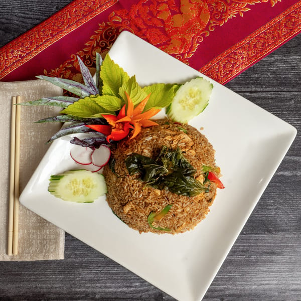 Thai fried rice with basil, cucumber, radish, and chili garnish on a white plate with chopsticks.