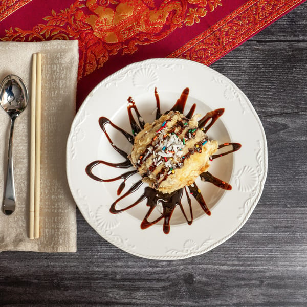 Fried ice cream with chocolate syrup and sprinkles on a white plate with chopsticks and spoon.