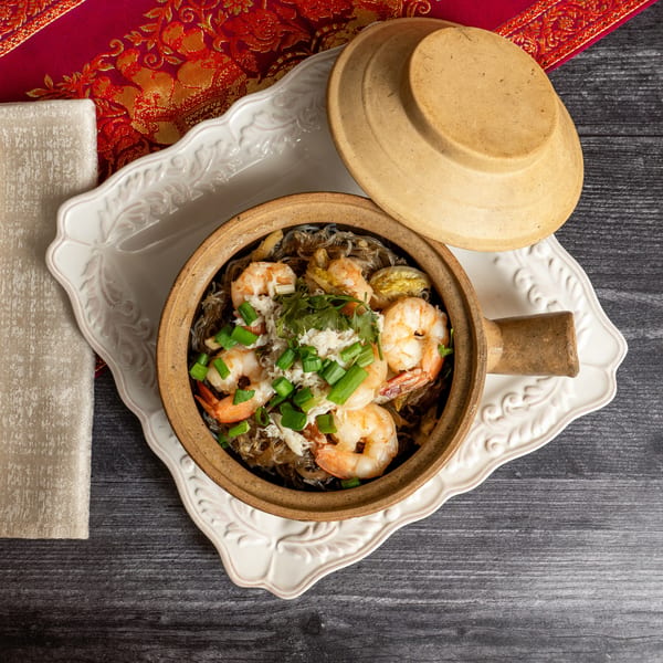 Clay pot with glass noodles, shrimp, crab meat, and green onions, with chopsticks and red fabric.